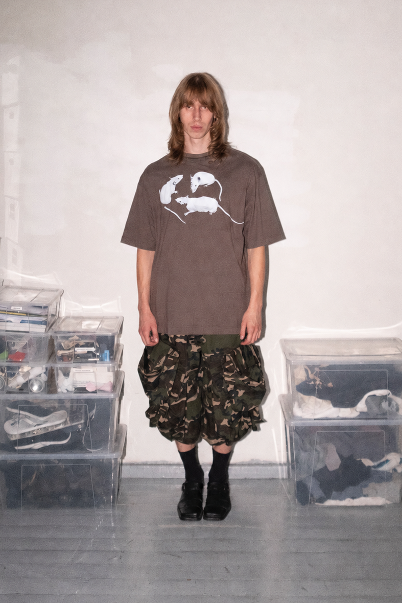 A person stands in a room wearing a brown oversized graphic T-shirt, camouflage ruffled shorts, black socks, and black shoes. Clear storage bins filled with items are stacked on both sides against a plain light wall.