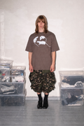 A person stands in a room wearing a brown oversized graphic T-shirt, camouflage ruffled shorts, black socks, and black shoes. Clear storage bins filled with items are stacked on both sides against a plain light wall.