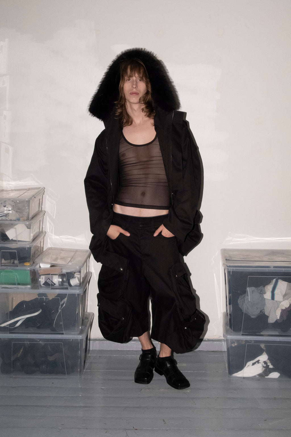 A model stands in a room wearing a black hooded jacket with a dark sheer top and black pants. The room has a white wall, gray floor, and clear plastic storage boxes filled with items on both sides.