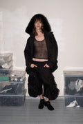 A model stands in a room wearing a black hooded jacket with a dark sheer top and black pants. The room has a white wall, gray floor, and clear plastic storage boxes filled with items on both sides.