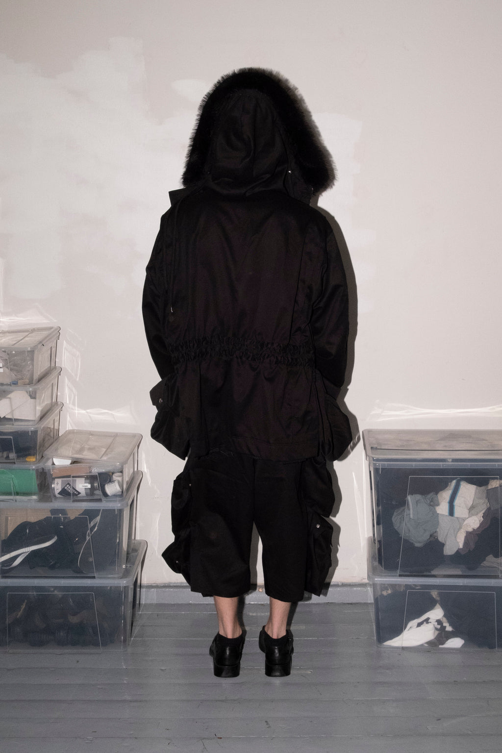 A person viewed from behind wearing a black hooded jacket with a dark fur-trimmed hood, black cropped pants, and black high heels, standing in a room with gray floors and white walls. Clear plastic storage bins are stacked on both sides.