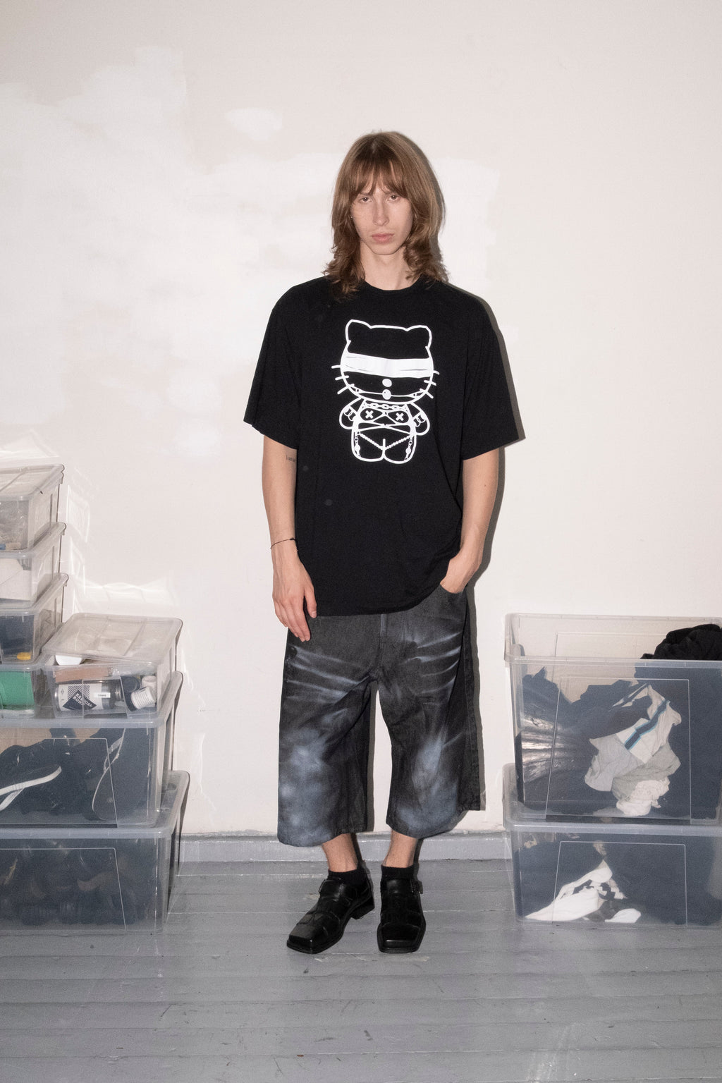 A person stands in a studio-like room wearing an oversized black graphic T-shirt with a white cartoon-style print, dark gray cropped pants, and black shoes. Clear storage bins and a white wall are visible in the background.
