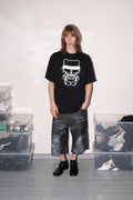 A person stands in a studio-like room wearing an oversized black graphic T-shirt with a white cartoon-style print, dark gray cropped pants, and black shoes. Clear storage bins and a white wall are visible in the background.