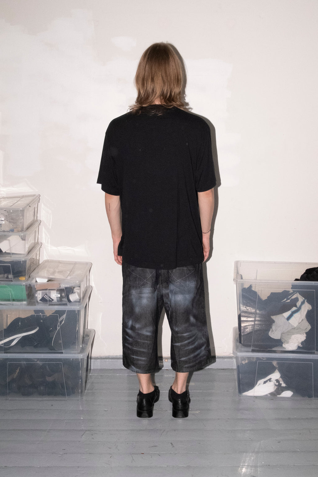 A person viewed from behind wearing a black oversized short-sleeve t-shirt and dark gray washed cropped pants, standing in a room with white walls and gray floor, surrounded by clear plastic storage bins containing clothing.