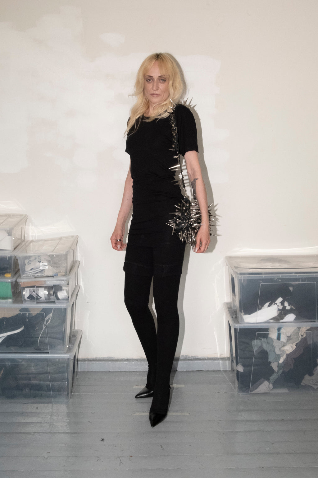 A woman stands in a studio wearing a black short-sleeve top with metallic spikes down the side, black pants, and black pointed shoes. She is posed in front of a white wall with clear storage bins on the floor.