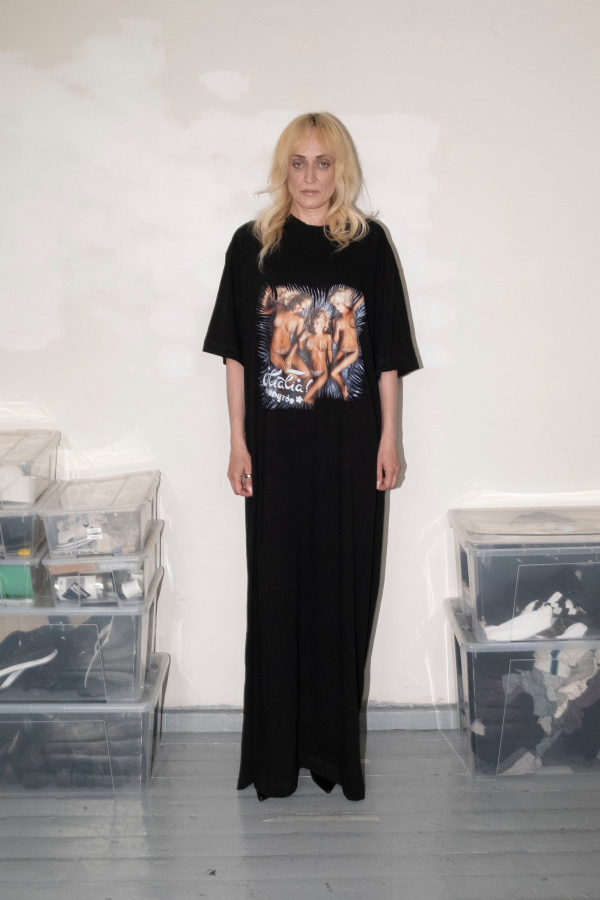 A woman stands in a room wearing a black maxi dress with a large graphic print on the front. The room has a white wall, gray floor, and clear plastic storage bins on both sides.