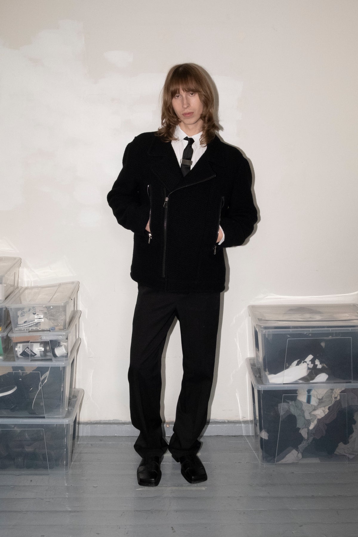 A person stands in a room wearing a black asymmetrical zip jacket over a white collared shirt with a black tie, paired with black trousers and black shoes. Clear plastic storage bins filled with clothing are stacked on both sides against a plain off-white wall.