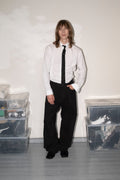 A person stands in a room wearing a white long-sleeve button-up shirt, black tie, and loose black trousers. Clear plastic storage bins filled with clothing are stacked on both sides against a plain off-white wall and gray floor.