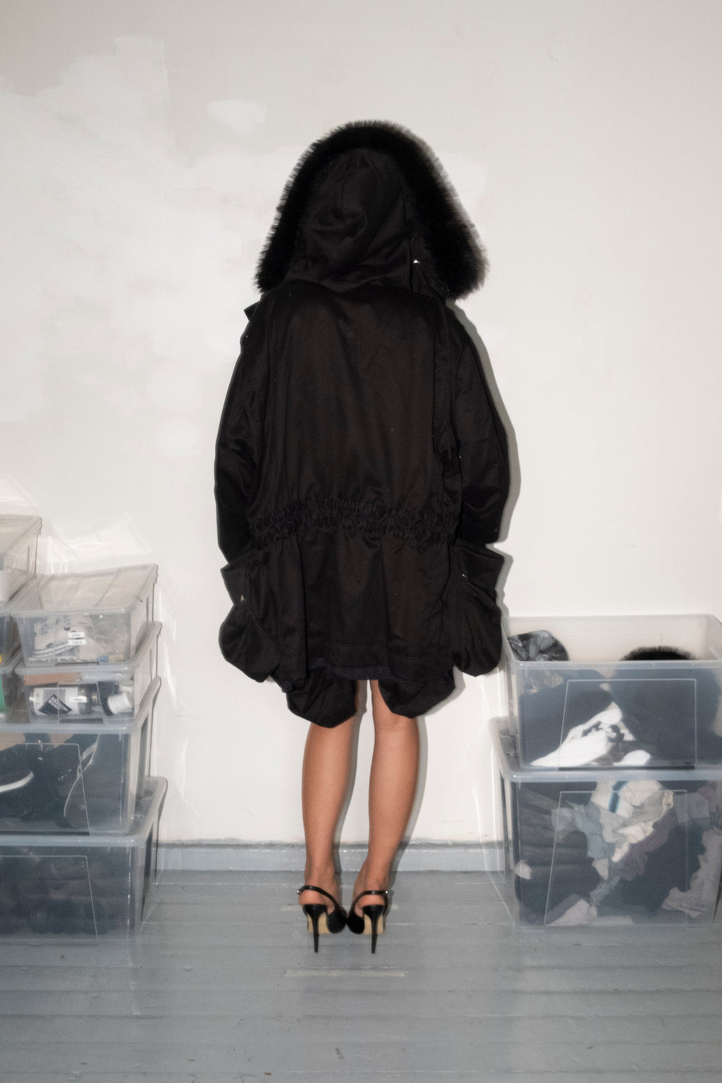 A person viewed from behind wearing a black hooded jacket with a dark fur-trimmed hood, standing in a room with white walls and gray floor, with clear plastic storage bins on both sides.