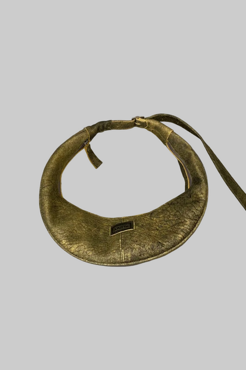 A vintage gold-toned metal collar or neckpiece with a small rectangular black label, displayed on a distressed white wooden surface.