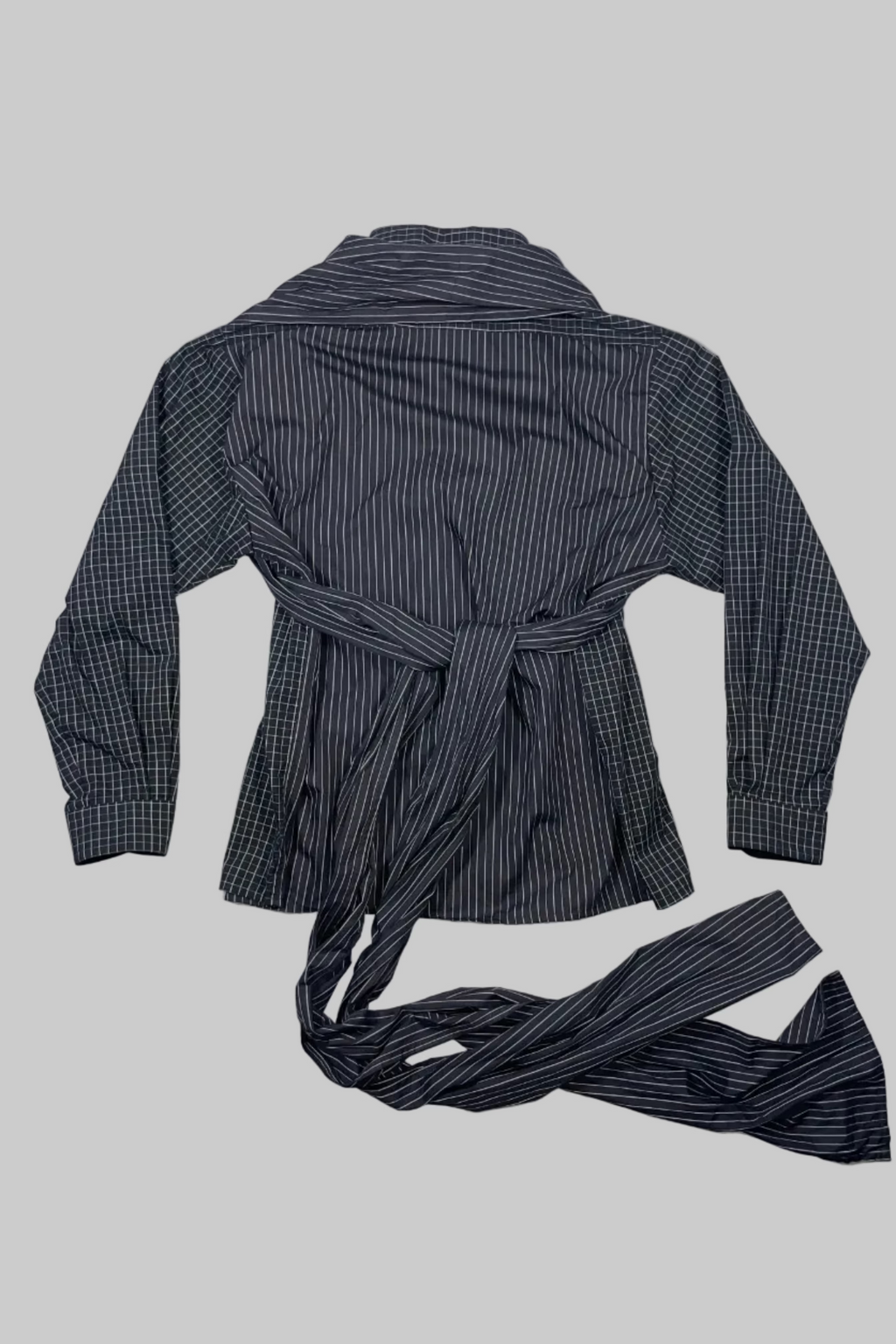 A long-sleeve belted shirt jacket laid flat on a white painted wood plank background. The garment features a dark gray and black striped body with a subtle plaid pattern on the sleeves and collar.