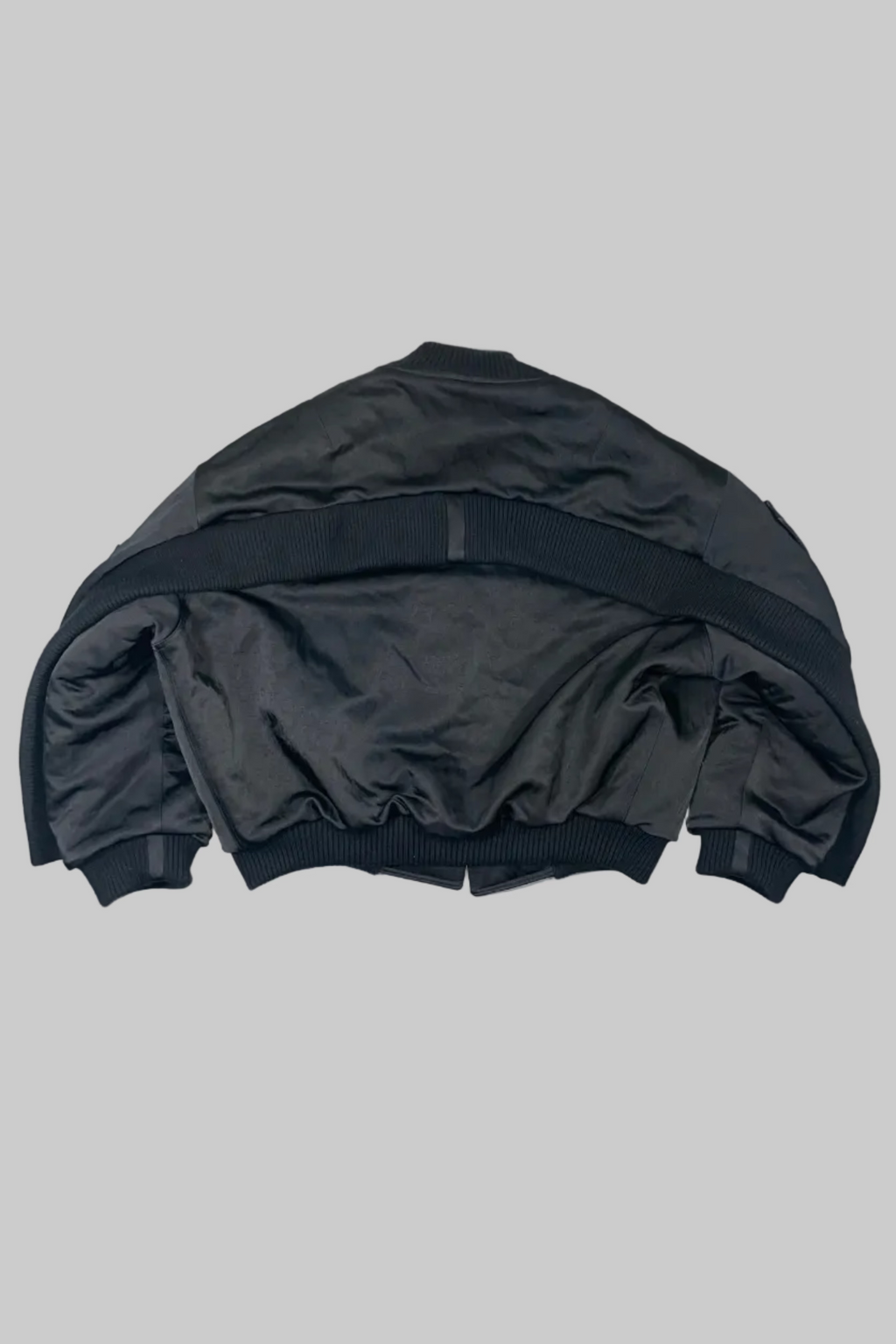 A black bomber jacket laid flat on a white painted wooden plank surface, showing the back view with ribbed collar, cuffs, and hem.