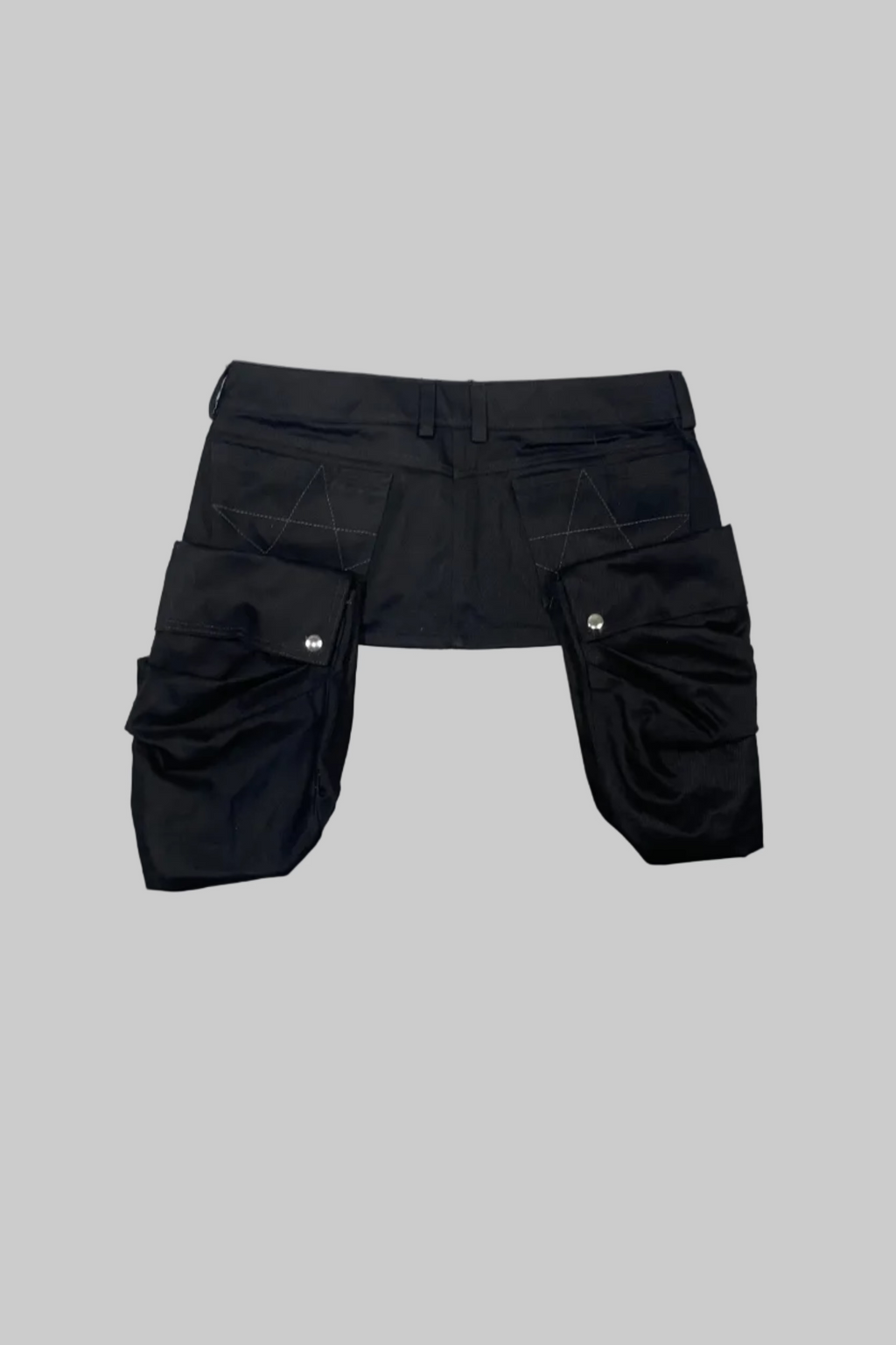 Black cargo shorts with a unique asymmetrical design, featuring a single front pocket on the right leg and a large multi-pocket cluster on the left leg, laid flat on a white wooden floor.