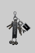 A metal keychain set with multiple keys, a rectangular black tag engraved with 'ESTAR', a small square black tag, and three silver bells, resting on a scratched light gray surface.