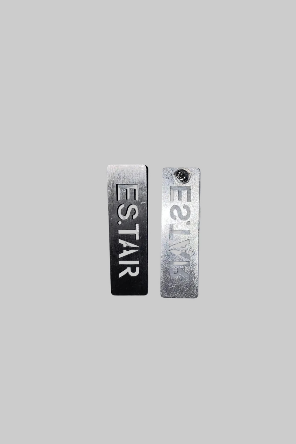 Two rectangular metal nameplates reading 'ES.TAR' in reverse, one black and one silver, mounted on a scratched gray metal surface.