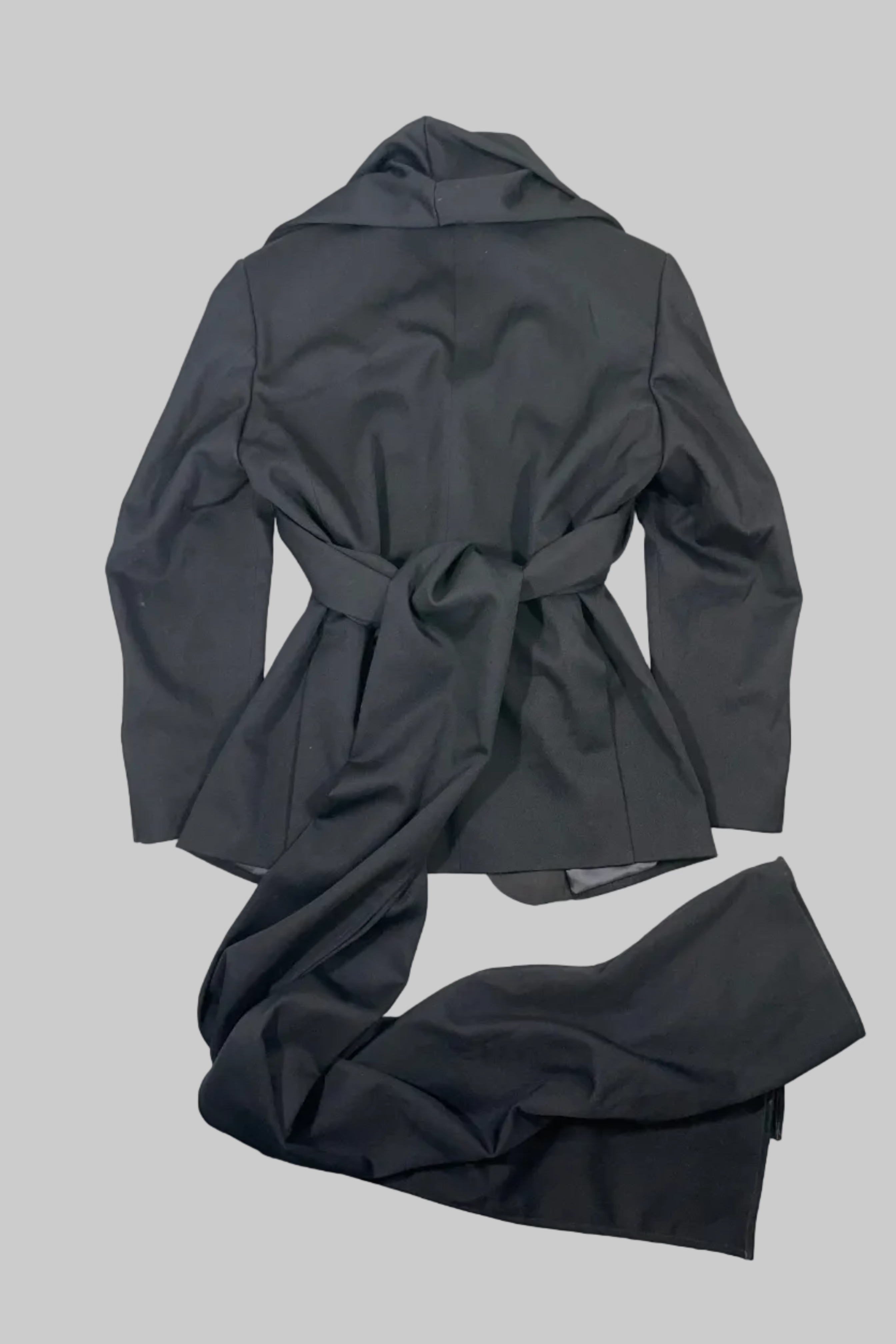 A dark gray belted coat or jacket laid flat on a white wooden plank surface, with matching dark gray trousers draped in front.
