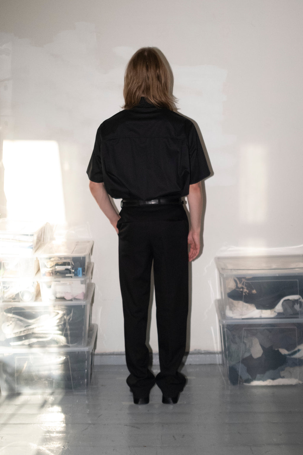 A person viewed from behind wearing a black short-sleeve collared shirt and black trousers, standing in a bright studio or room with white walls and floor, with clear plastic storage bins on both sides.