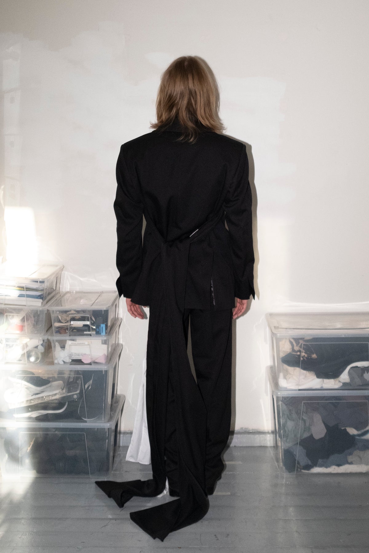 A model viewed from behind wearing a black oversized belted blazer and matching wide-leg trousers, standing in a minimalist studio with white walls, gray floors, and clear storage bins.