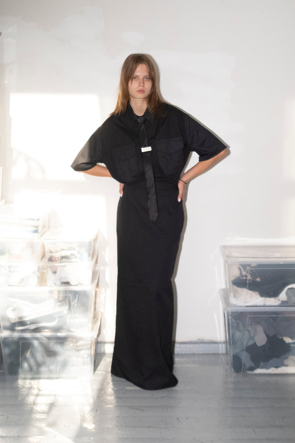 A model stands in a minimalist studio wearing a black long-sleeved maxi dress with a wide collar and a matching black belt. The room has a white wall, gray floor, and clear plastic storage bins on either side.