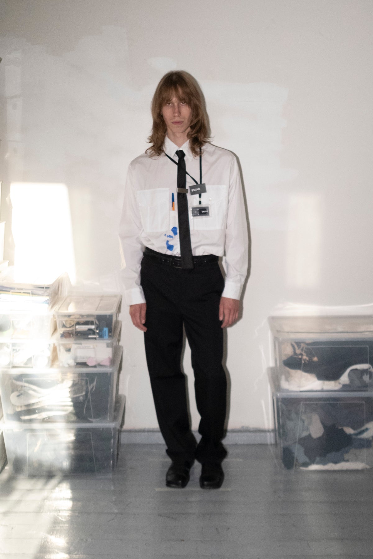 A person stands in a room wearing a white long-sleeve button-up shirt with a black necktie, black trousers, and black shoes. Clear plastic storage bins are stacked against a white wall on both sides.