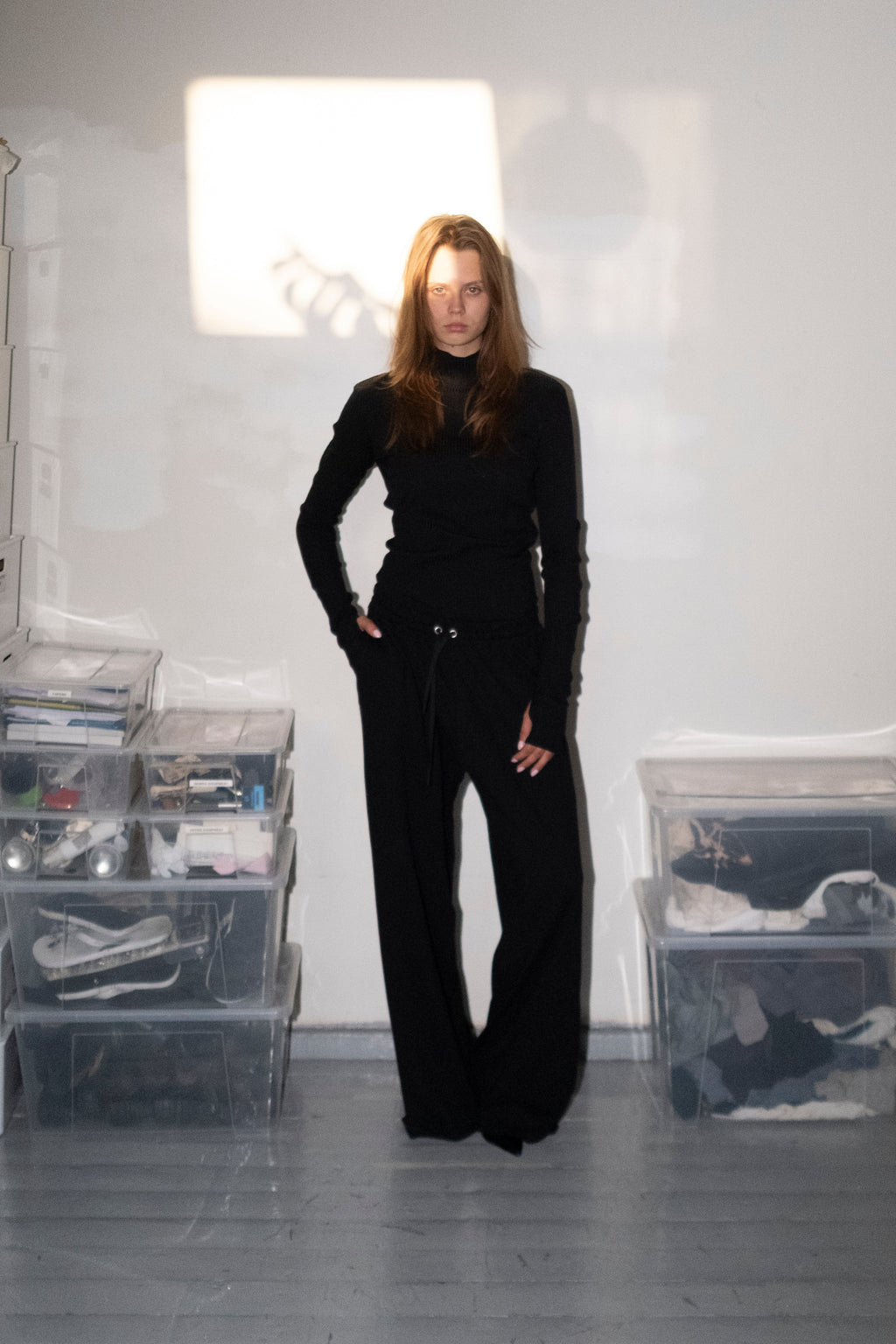 A woman with long light brown hair stands in a minimalist room wearing a black long-sleeve top and matching black wide-leg pants. She is positioned in front of a white wall with a bright light reflection, flanked by clear plastic storage bins on both sides.