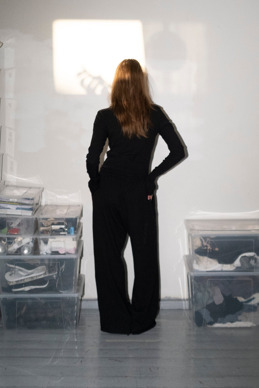 A woman stands with her back to the camera in a minimalist room, wearing a black long-sleeve top and matching wide-leg pants. Clear plastic storage bins filled with clothes are stacked on both sides against a white wall with a bright light projection.