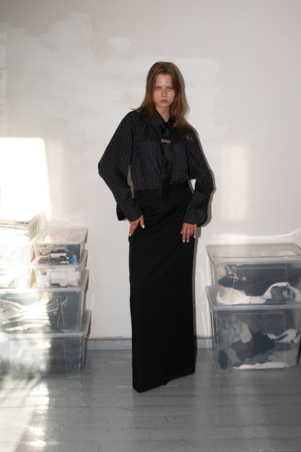 A model stands in a minimalist room with white walls and gray floors, wearing a black long-sleeved top with a wide collar and a long black skirt. Clear plastic storage bins are stacked on both sides of the room.
