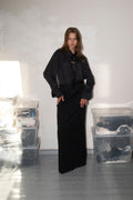 A model stands in a minimalist room with white walls and gray floors, wearing a black long-sleeved top with a wide collar and a long black skirt. Clear plastic storage bins are stacked on both sides of the room.