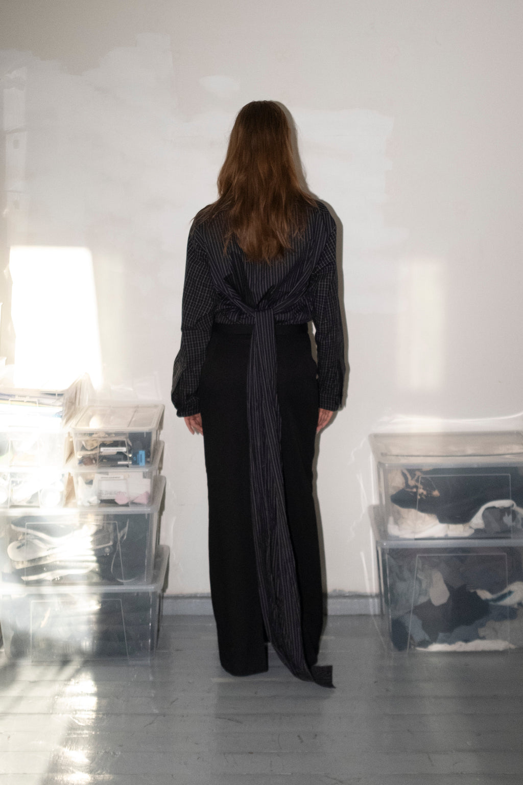 A woman stands with her back to the camera in a minimalist studio space, wearing a dark gray long-sleeve wrap-style top and black wide-leg trousers. Clear plastic storage bins are stacked on the floor on both sides against a white wall.