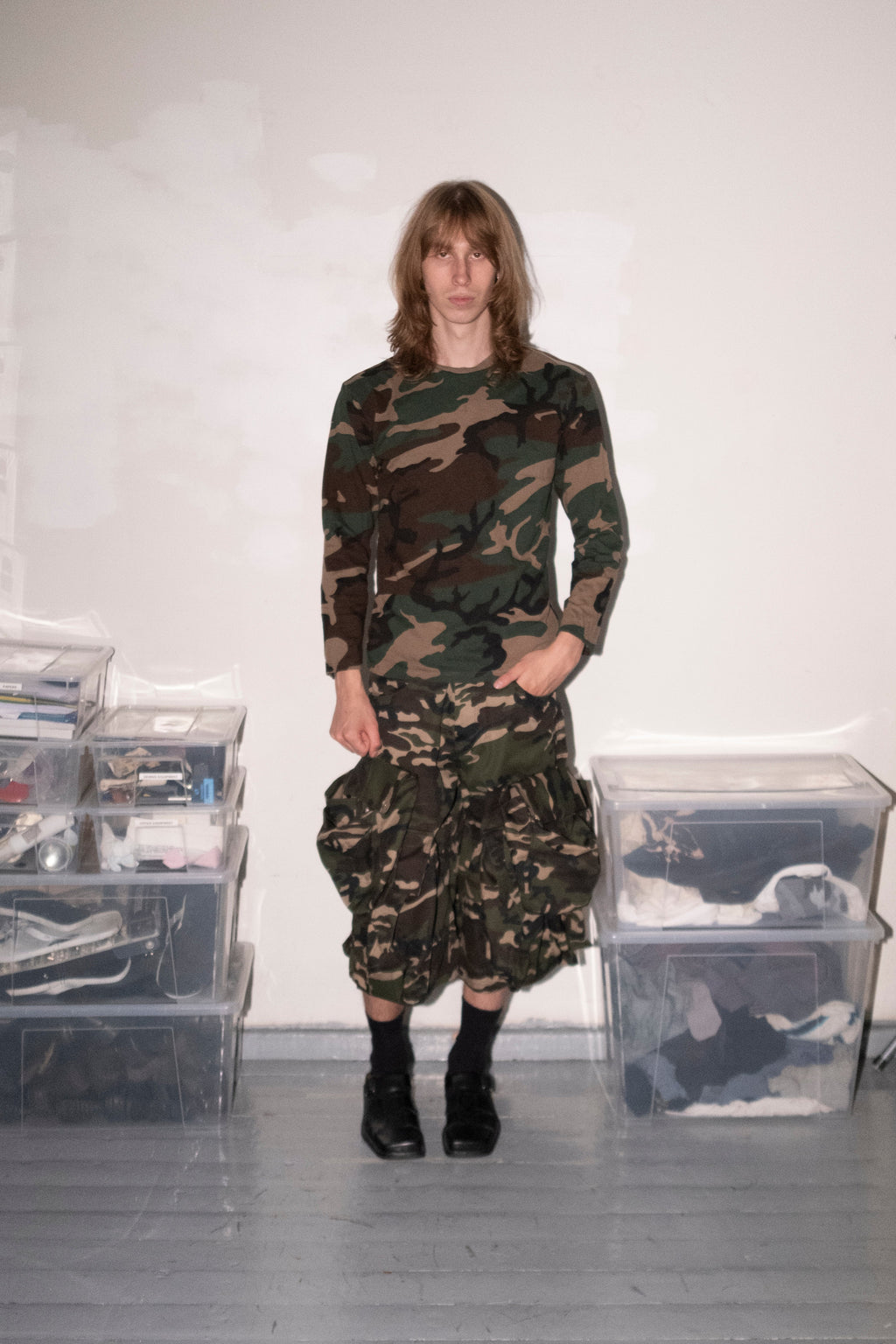 A person stands in a room wearing a camouflage long-sleeve top and matching camouflage skirt or pants, paired with black socks and black shoes. Clear plastic storage bins are stacked on both sides against a plain white wall.