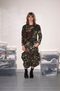 A person stands in a room wearing a camouflage long-sleeve top and matching camouflage skirt or pants, paired with black socks and black shoes. Clear plastic storage bins are stacked on both sides against a plain white wall.