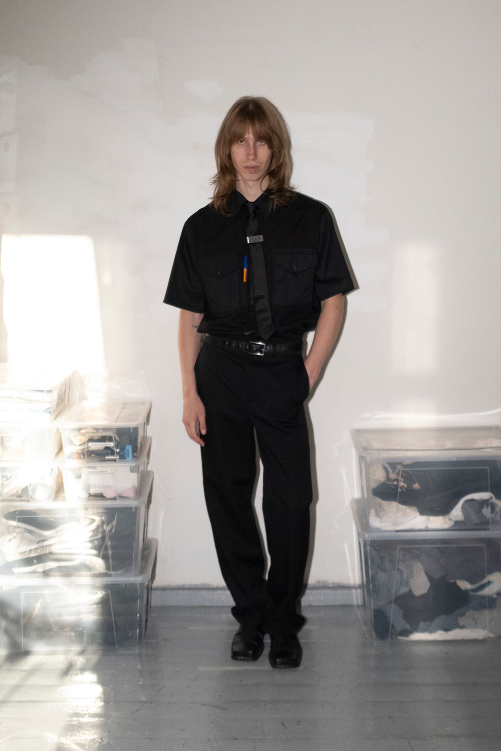A model stands in a minimalist studio wearing a black short-sleeve button-up shirt, black trousers, and a black tie. The room has a white wall, gray floor, and clear plastic storage bins on either side.