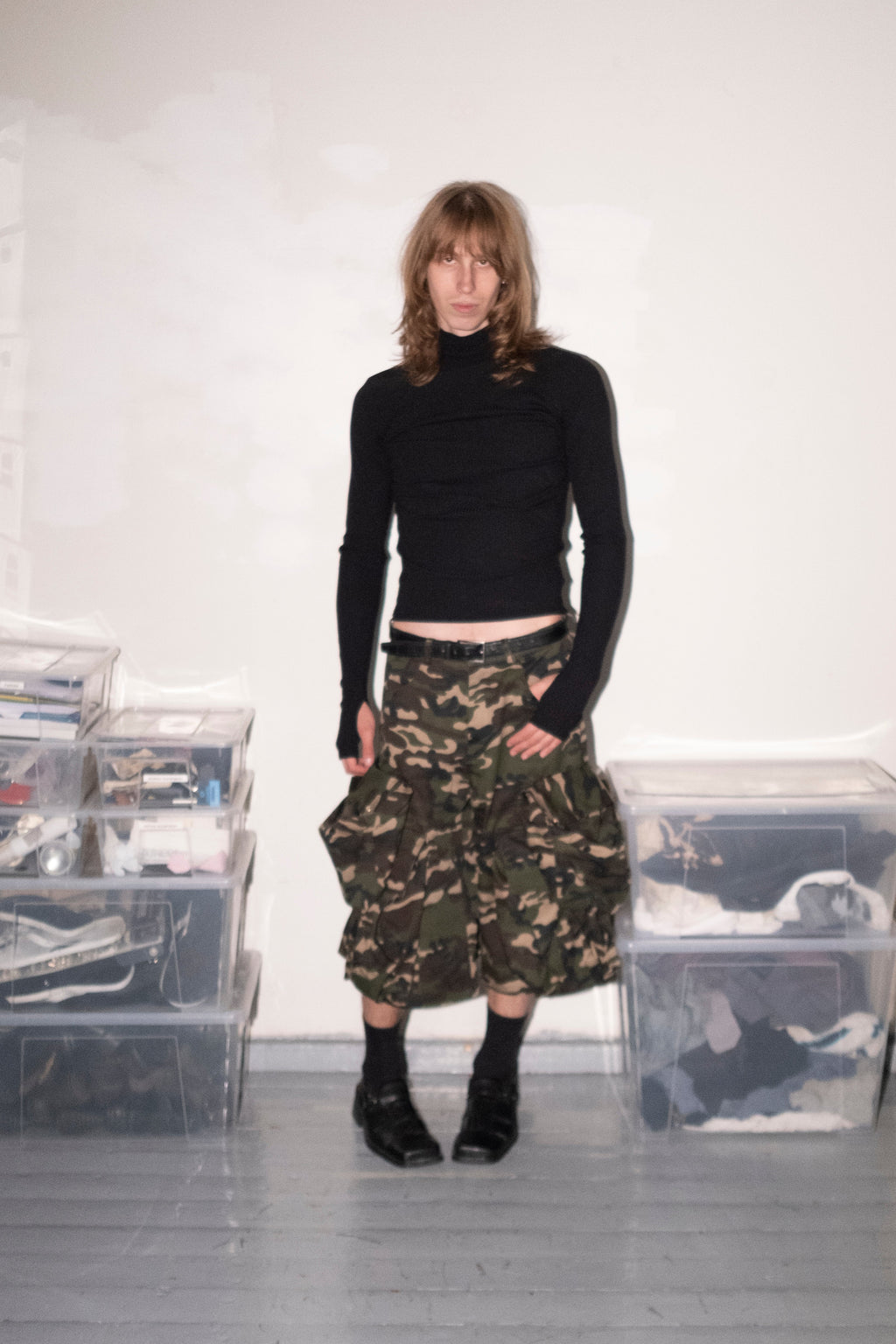 A model stands in a room wearing a black long-sleeve crop top and a high-waisted, voluminous camouflage skirt with black boots. Clear plastic storage bins filled with clothing are stacked on both sides against a white wall.