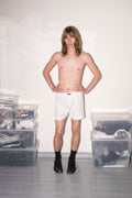 A shirtless young man with shoulder-length light brown hair stands with hands on hips wearing white boxer briefs, black socks, and black boots in a minimalist room with white walls and gray floors. Clear plastic storage bins are stacked on both sides.